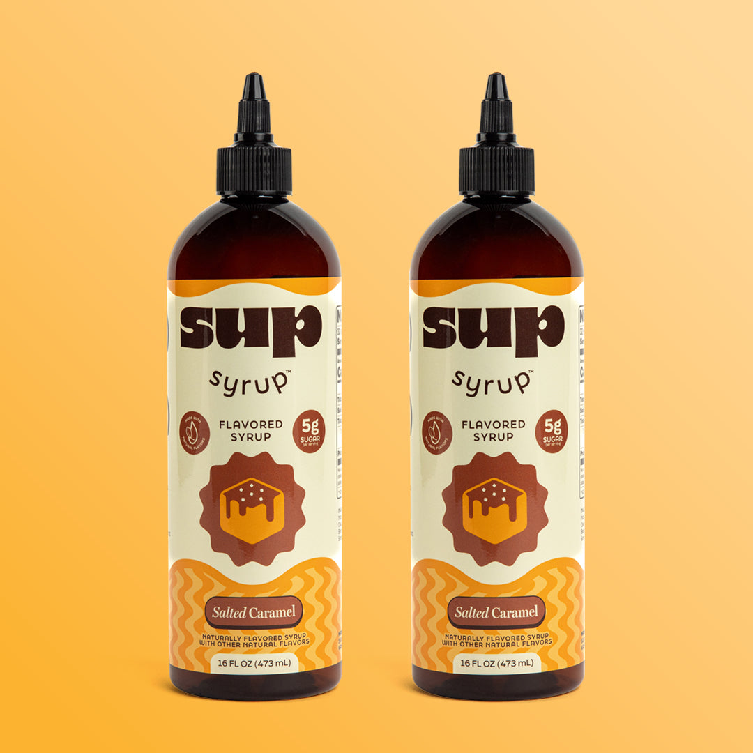 Two 16 fl oz amber bottles of Sup Syrup Salted Caramel flavored syrup with a black twist open cap, set against an orange background.