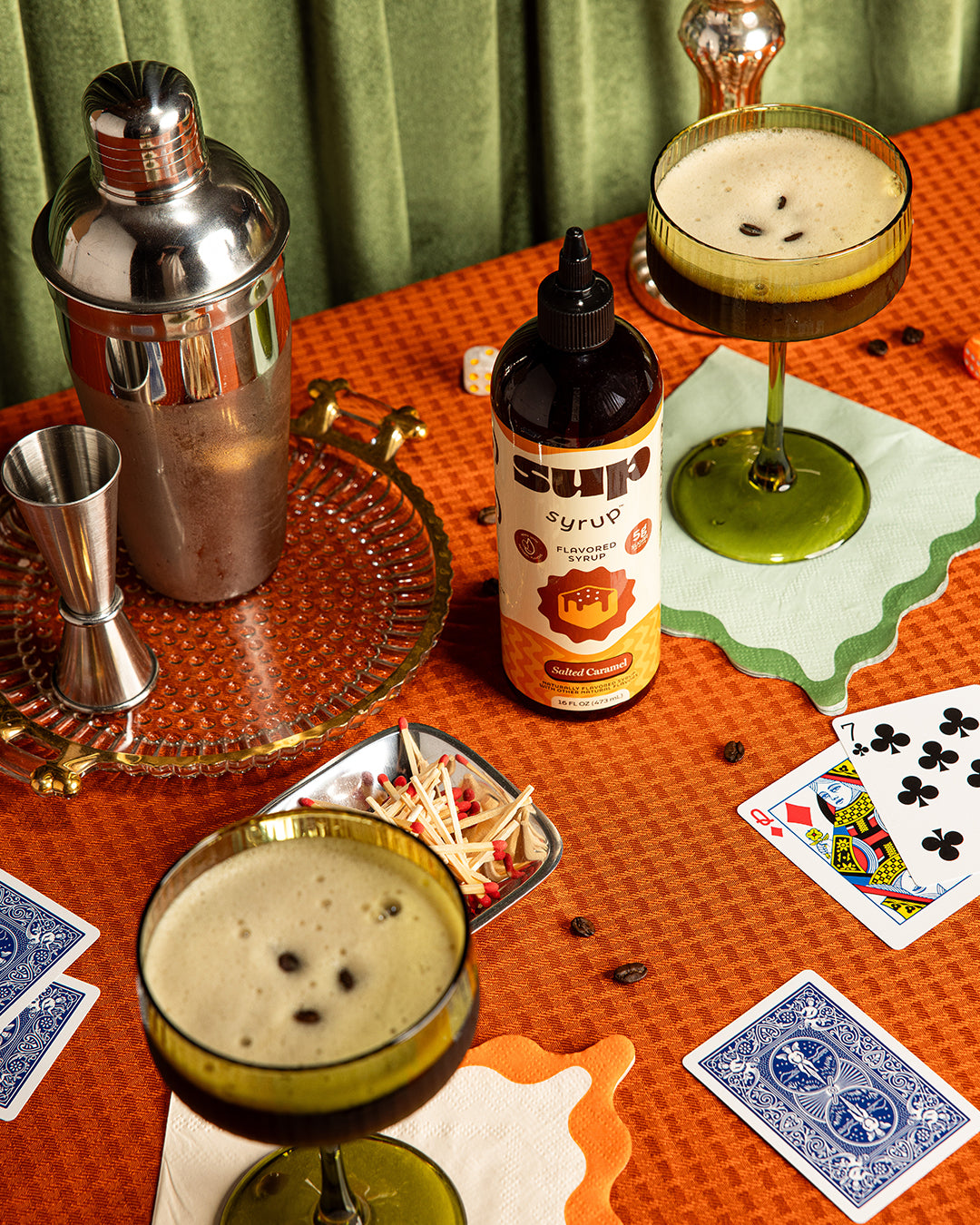 Cocktail shaker, Sup Syrup bottle, and cocktails on a table with playing cards.