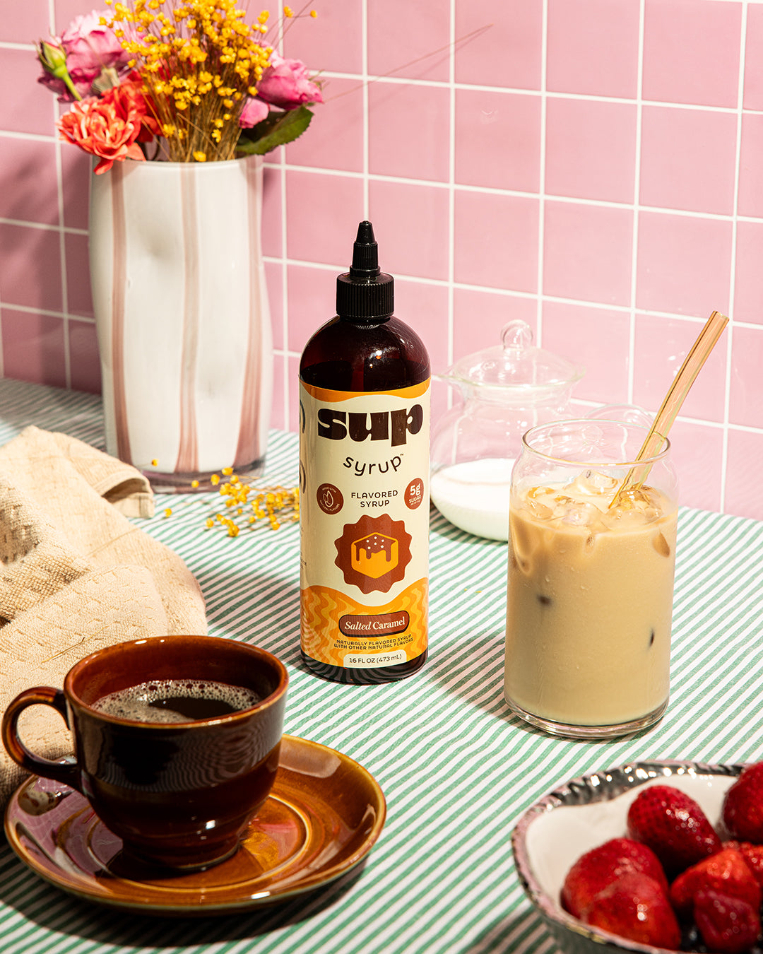 Table setting with a bottle of Sup Syrup, a cup of coffee, iced coffee, and strawberries against a pink tiled wall.