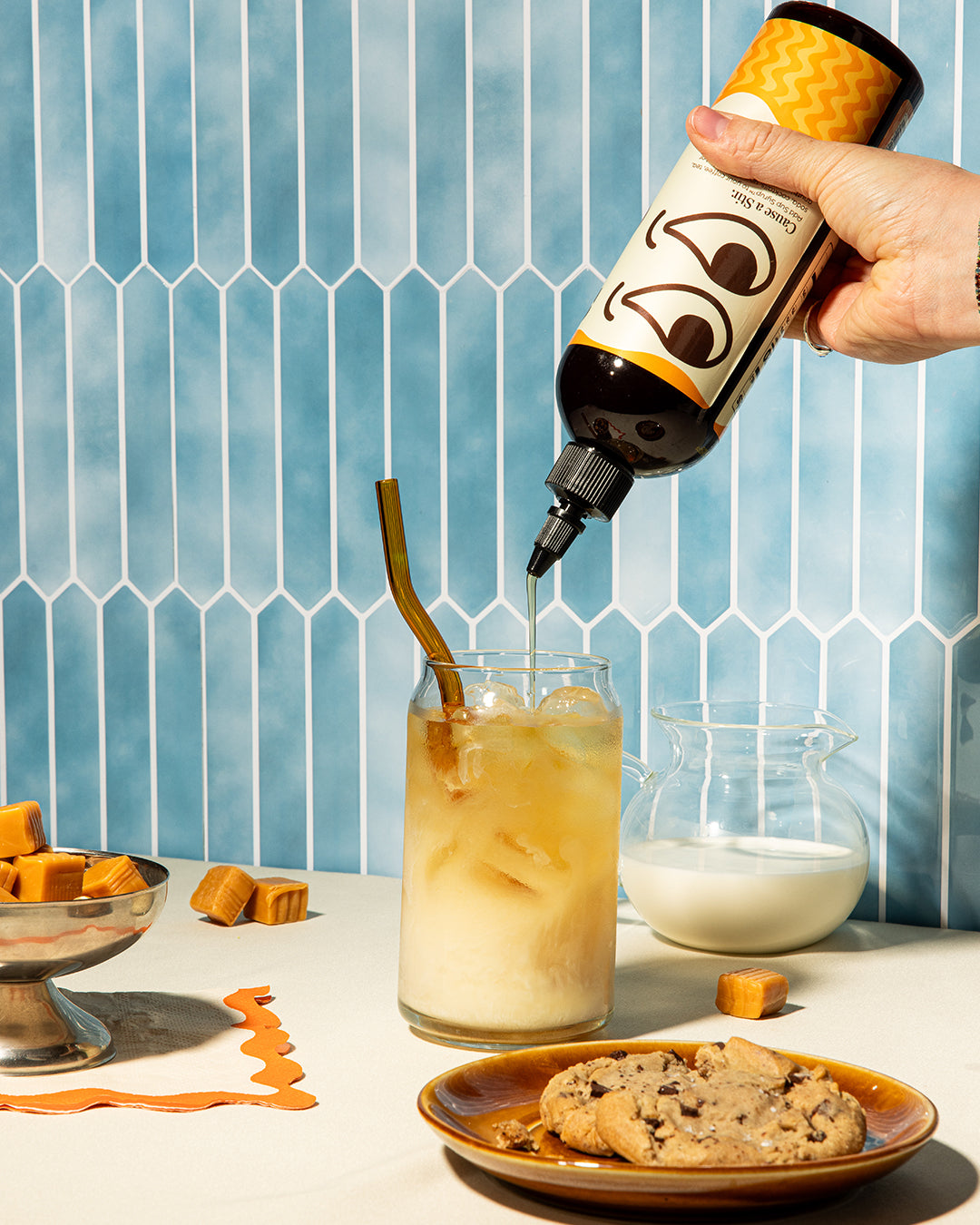 Hand pouring a bottle of Sup Syrup into a glass of dirty cream soda with ice and a straw, against a blue tiled wall.
