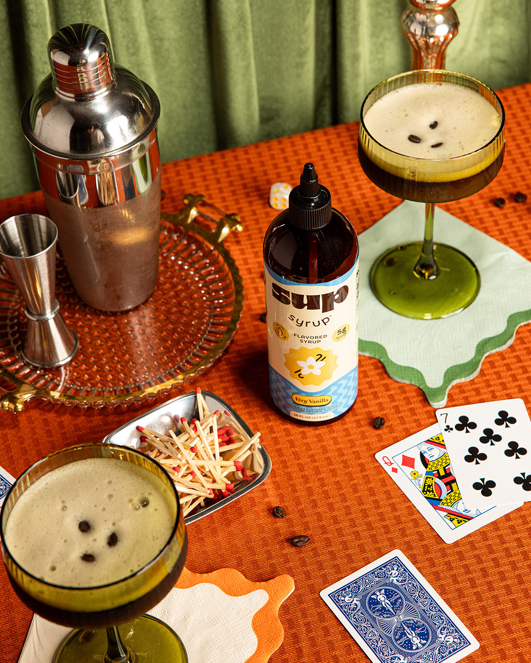 Cocktail shaker, glasses with espresso martinis, bottle of Very Vanilla Sup Syrup, and playing cards on a table.