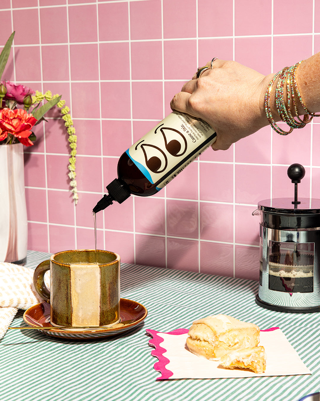 Hand pouring Very Vanilla Sup Syrup into a coffee cup with a pink tiled wall background.