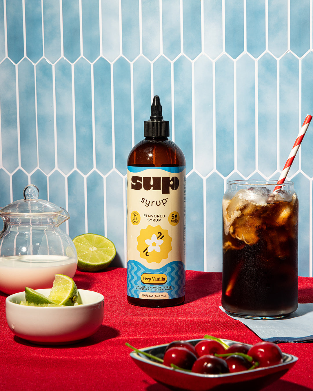 Very Vanilla Sup Syrup bottle with a glass of dirty soda on a red surface against a blue tiled wall.