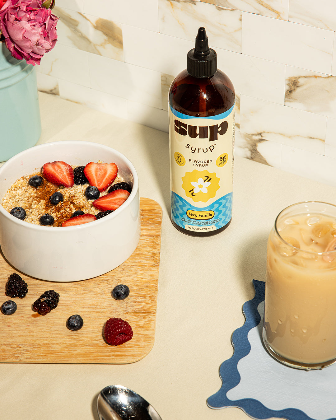 Bottle of Very Vanilla Sup Syrup next to a bowl of oatmeal and fruit and a glass of iced coffee on a table.